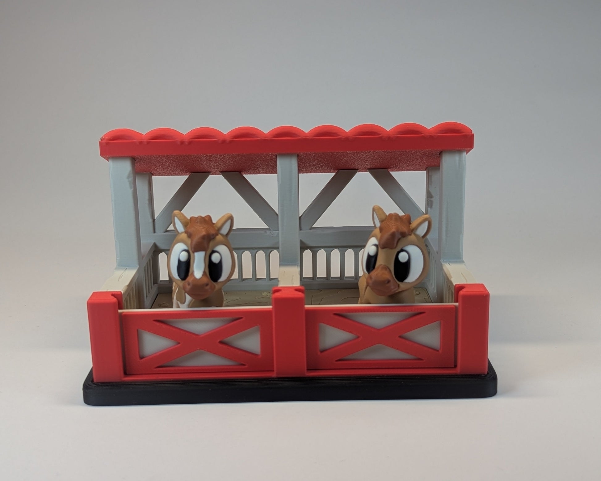 Two toy horses in a red and gray stable on a light gray background